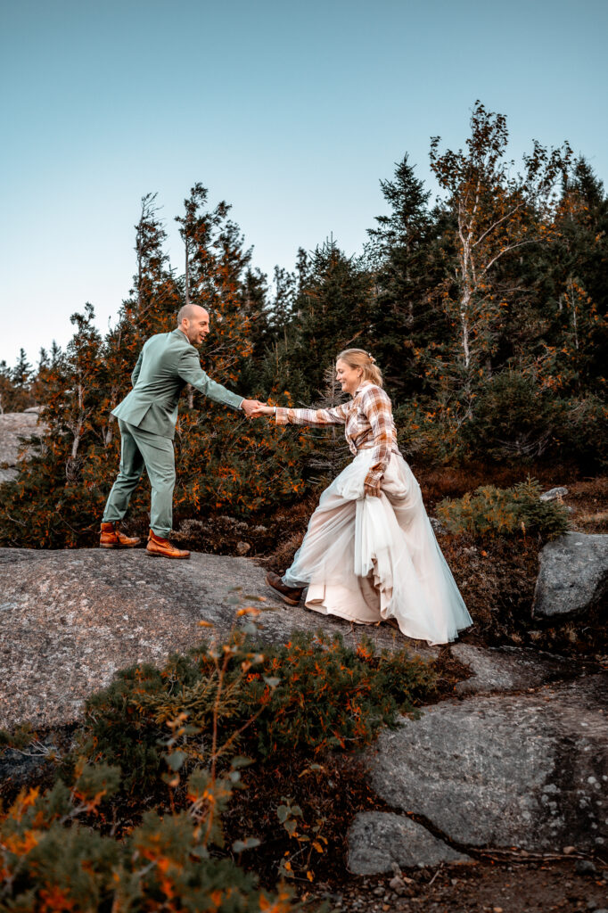 Groom takes brides hand and helps her up a rock face.