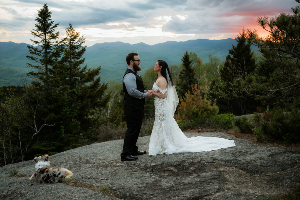 A couple says "I Do" on top of a mountain at sunset on a rocky summit while they have their australian shepherd laying down next to them with mountains in the background. 
