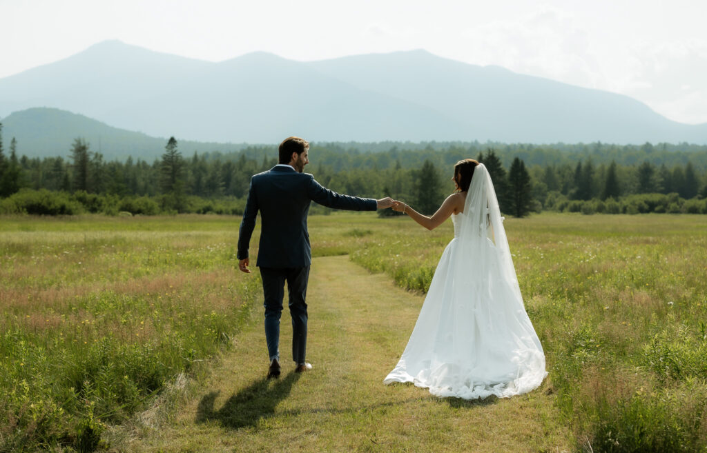 A couple holds hands and walks through a mountain field with mountains in the background while they are wearing a white wedding dress and a dark blue tux. 