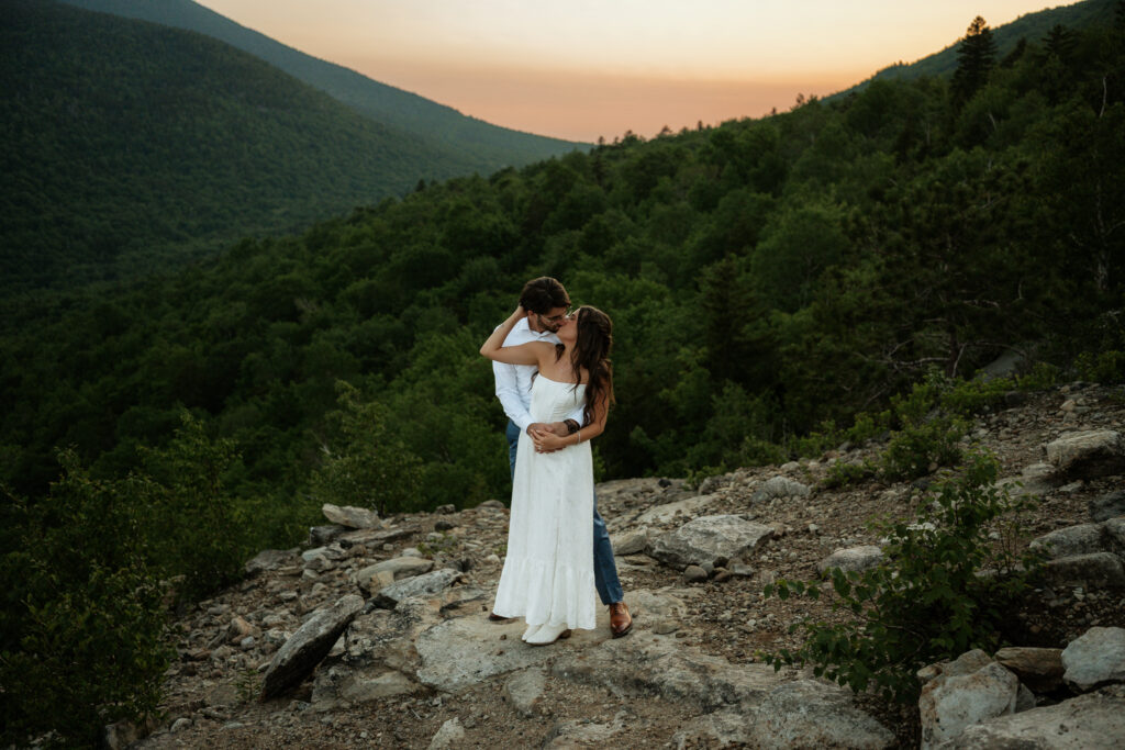 A couple in wedding attire hold eachother with the groom behind the bride, they are on top of a mountain summit with the sunset in the background and the valley connecting where the couple is standing. 