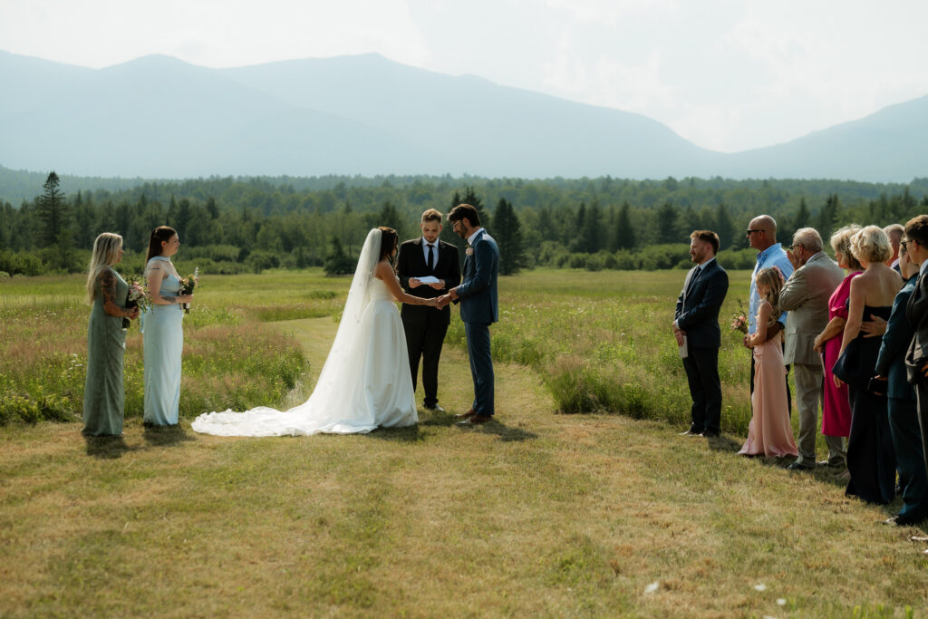 A couple who found a location for where to elope in the Adirondacks, in a field with family and mountains in the background on a sunny day. 