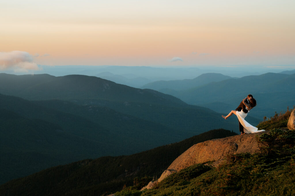 A couple does a dip kiss in wedding attire on top of an Adirondack High Peak with mountains and a valley behind them at sunset. 