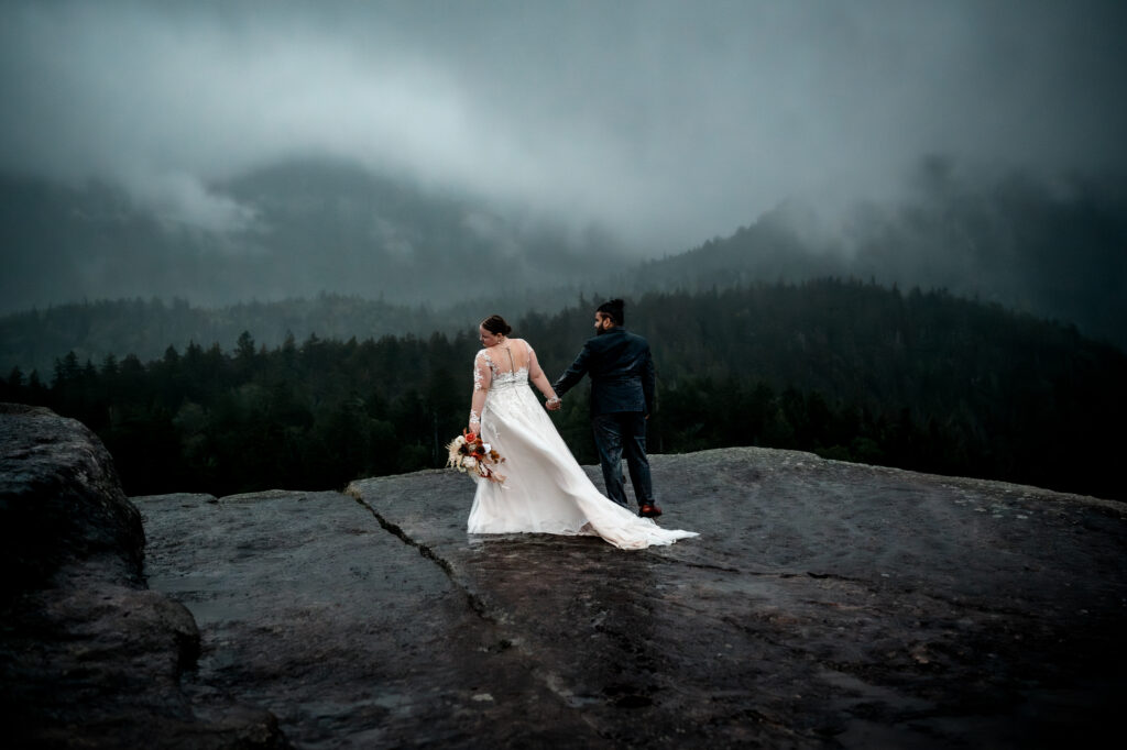 Couple holds hands on a misty and cloudy mountain peak while they look out at pine trees.