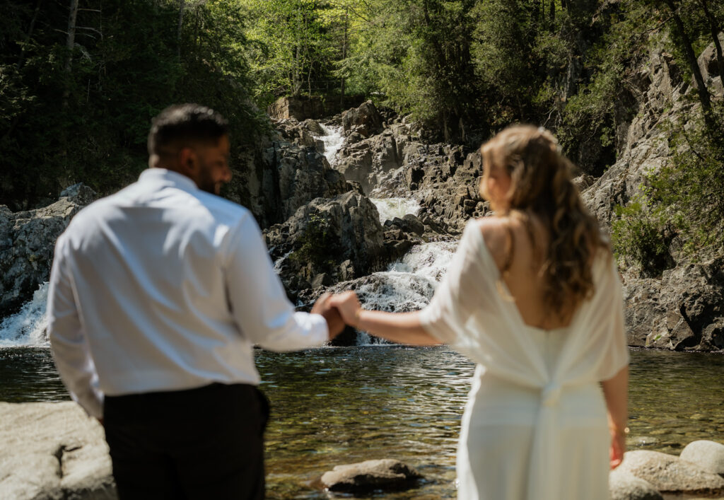 A couple stands at the base of a waterfall after getting married, they are holding hands looking at each other and out of focus while the waterfall in front of them is in focus and rushing. 