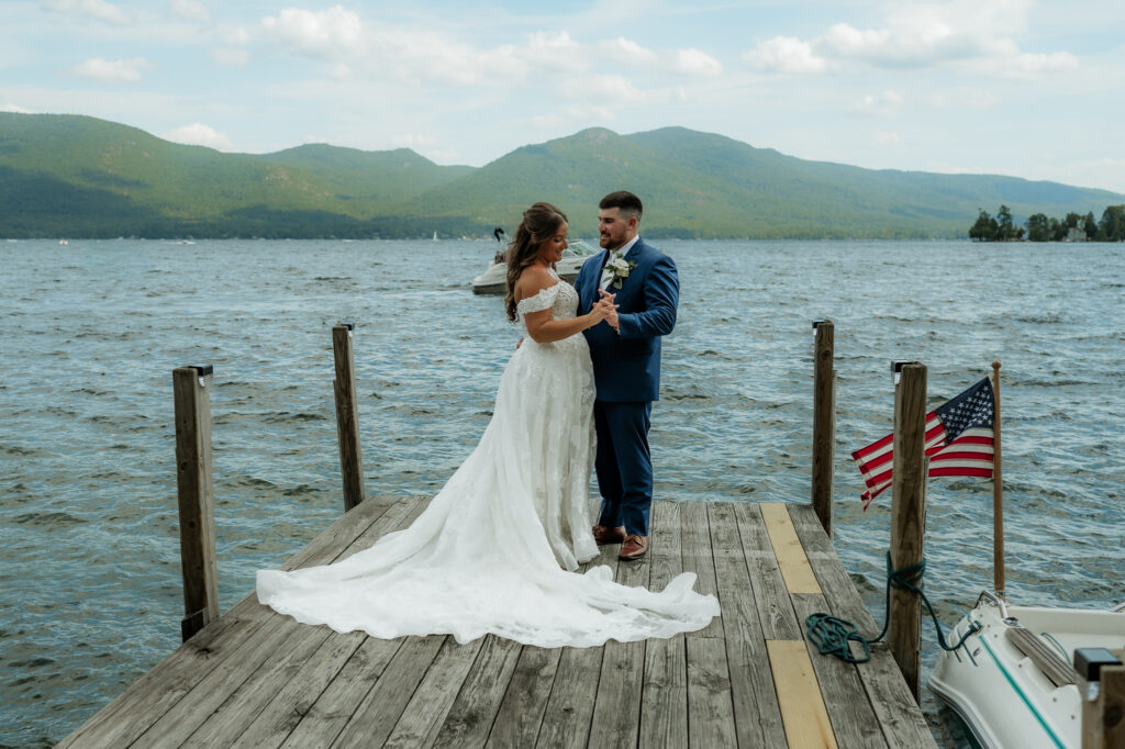 A couple stands on a dock in Lake George after they just got married in a blue tux and white dress with mountains and boats in the background. 