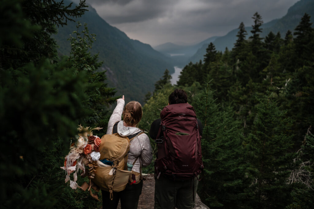 Couple points out where the sun is peaking out from behind the mountains on a cloudy day during their hike to their elopement. 