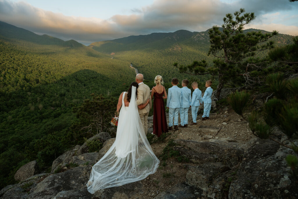 A bride and groom hold each other on a rocky summit in the Adirondacks looking away at a Valley of lush green mountains at sunrise while their five children who are dressed in blue and red stand next to them holding hands looking out at the mountains.