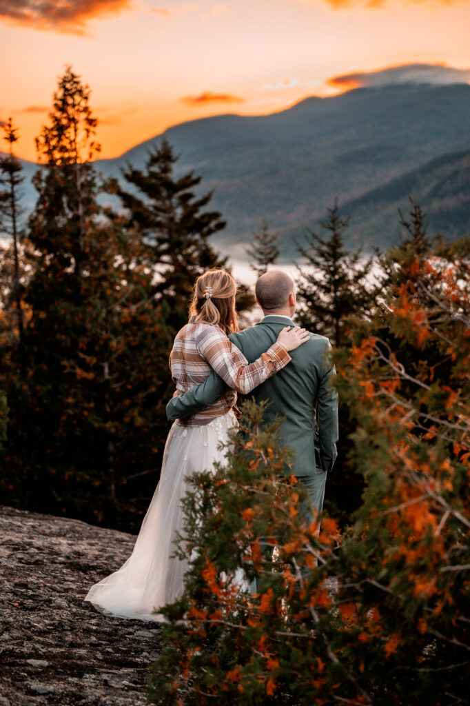 A couple looks out at the sunrise at mount jo in the adirondacks holding eachother.