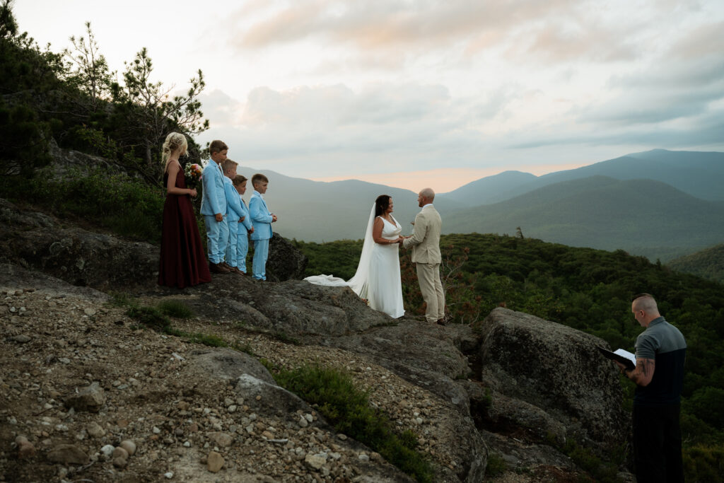 Sunrise elopement with kids in the Adirondack Mountains where the couple is standing on a rock ledge with their kids next to the them and they are in wedding attire with green mountains behind them and a sky with  cotton candy clouds.