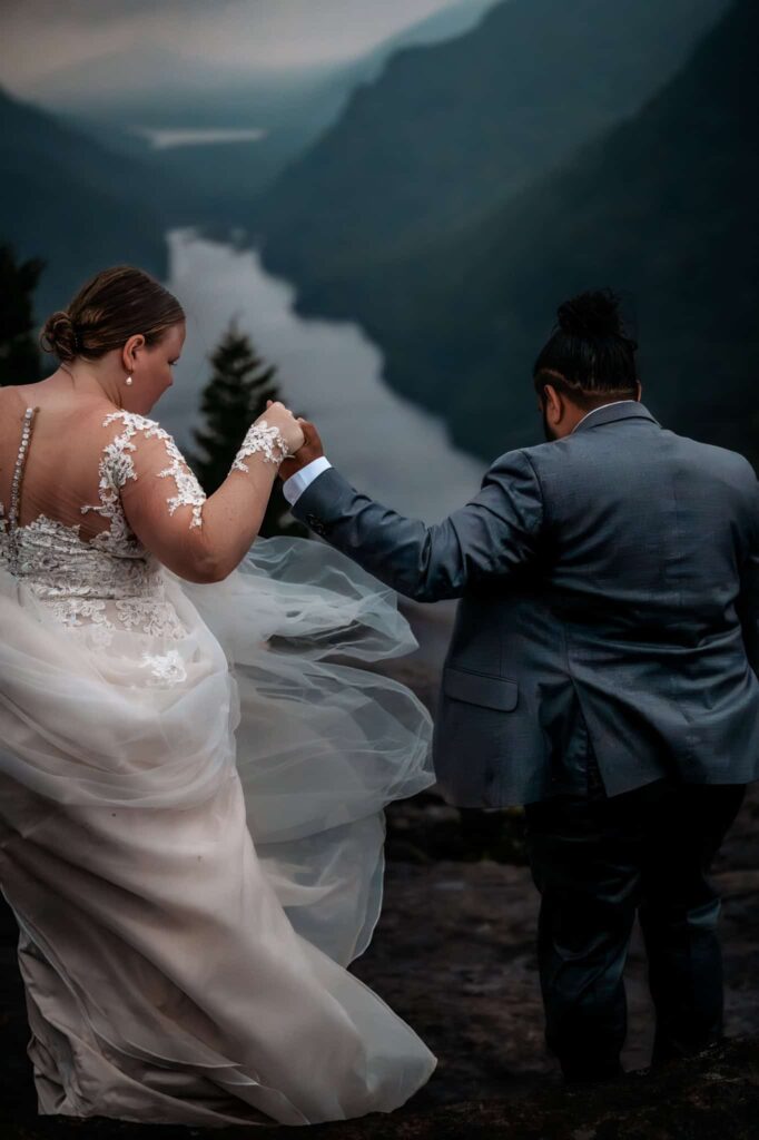 A couple holds hands as the groom guides the bride down a set of rocks on top of indian head summit in the adirondacks on a cloudy summer day.