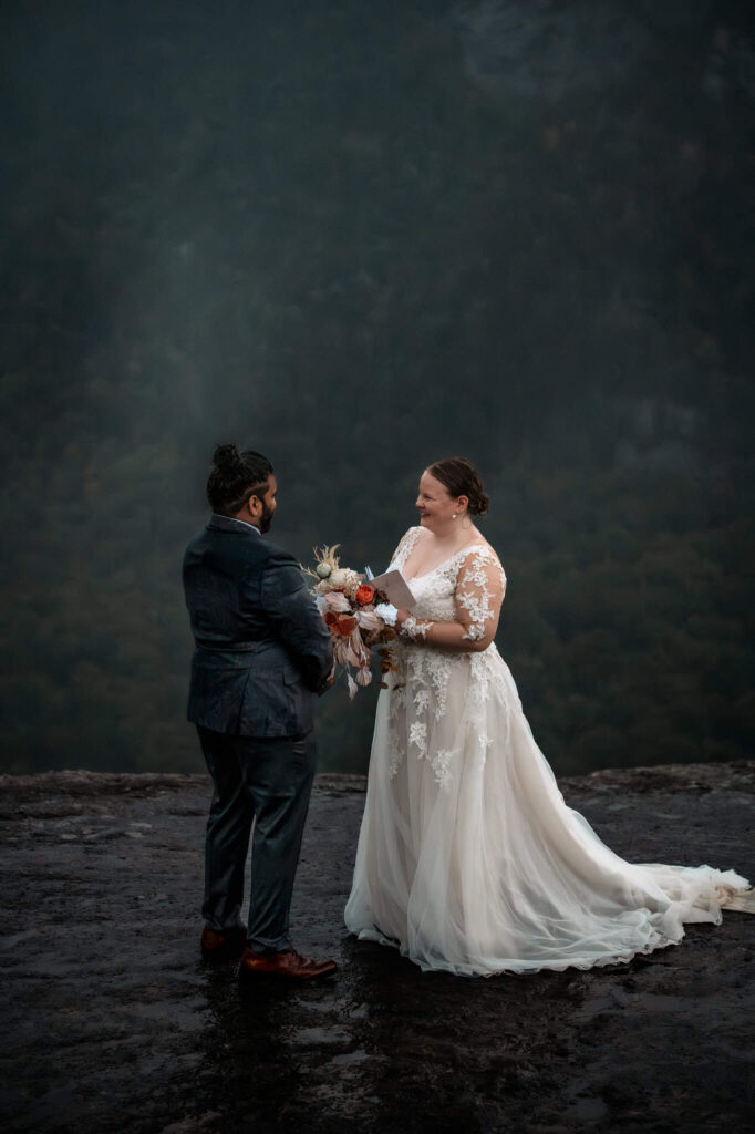 A couple exchanges vows on a mountaintop in a valley while it's raining.