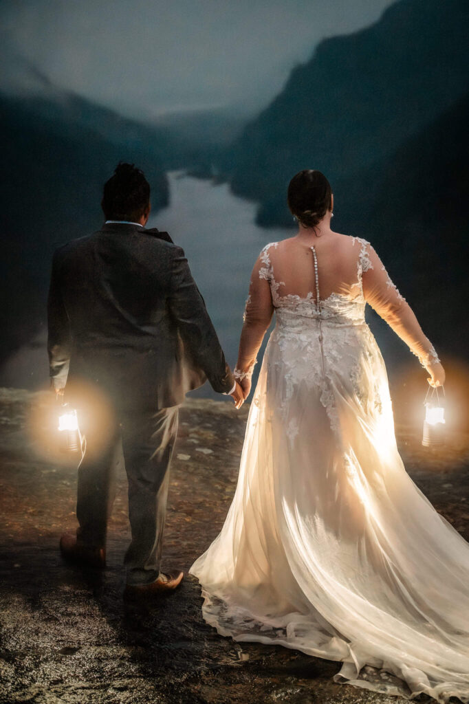 A couple holding lanterns looking out at ausable river at indian head summit, holding hands after eloping. 