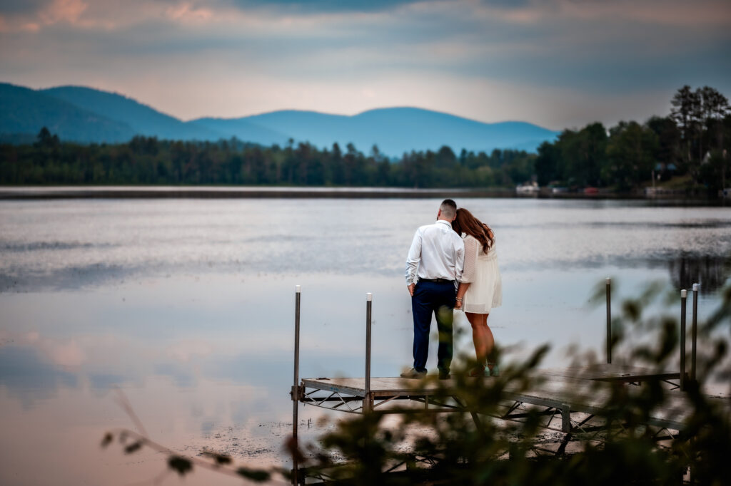 A couple looks out at a lake with a mountain backdrop on a dock. 