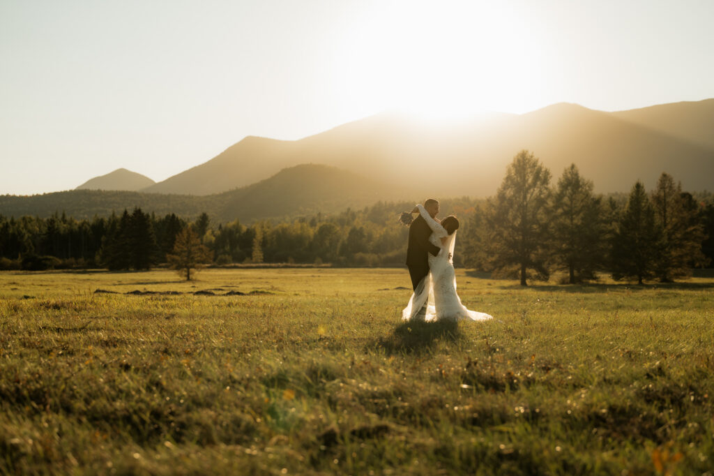 A couple holds onto each other in a field with mountains in the background at sunset in the fall with the sun setting over the mountains and they are in wedding attire with a bride and groom. 