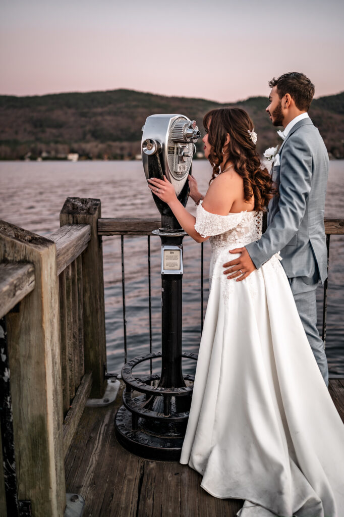 A couple who eloped looks through telescope at lake george on a dock. 