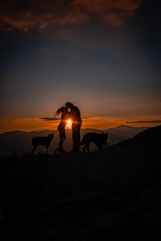 A silhouette couple kisses at sunset with the sun peeking through their bodies on a mountain summit with their dogs. 