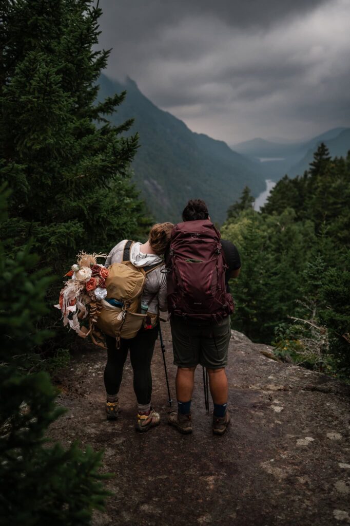 A couple hikes looks out at a mountain valley at Indian Head Mountain Summit with their hiking gear on and the bride is resting her head on the groom.
