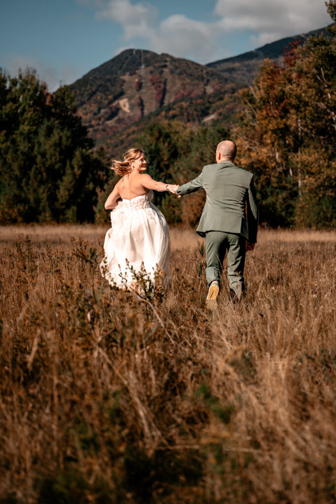 A couple chases each other in a field during fall in front of whiteface mountain after getting married.