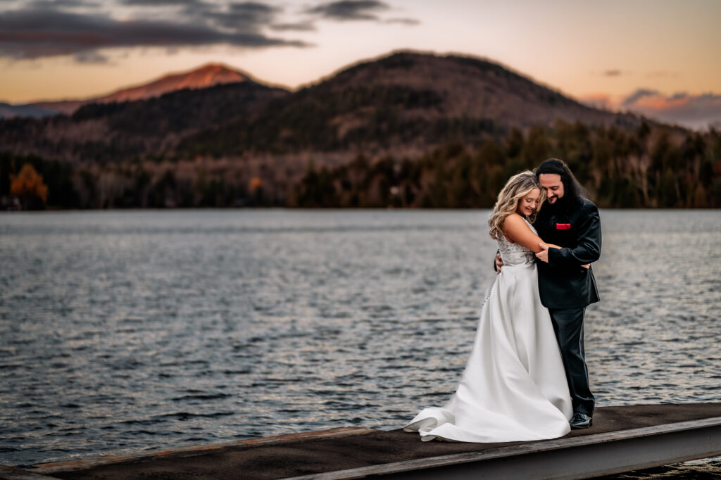 A couple stands on a dock while looking down at the water after getting married in front of mountains on mirror lake in lake placid.