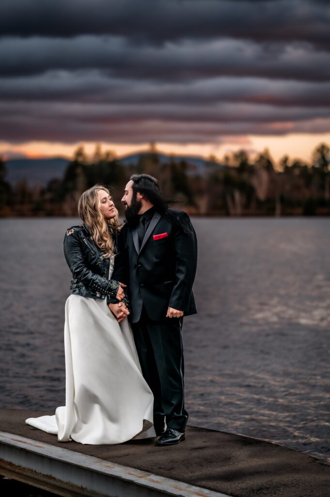a couple looks at eachother with passion while holding hands on a dock on mirror lake in lake placid agt sunset with colorful clouds behind them. 