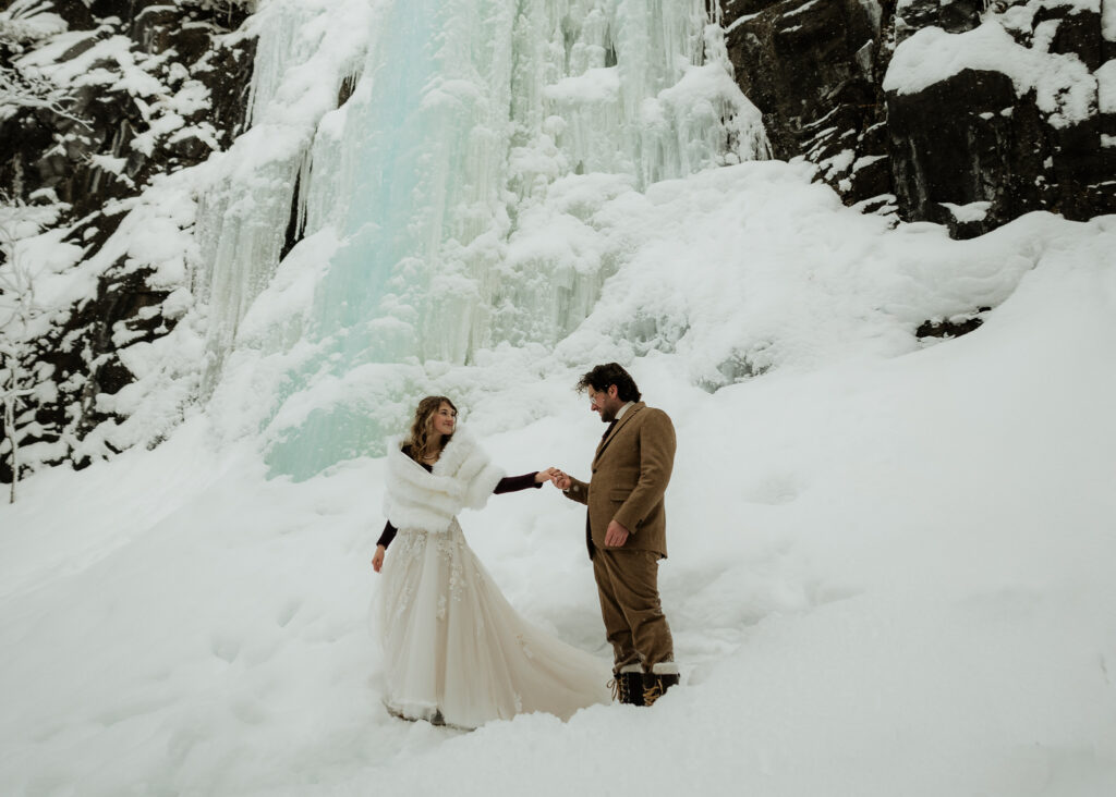 Winter Adirondack Elopement at a frozen waterfall in the snow with a couple who is standing at the base of the waterfall holding hands and the bride is in a white dress with a furry shaw and the groom is wearing a tan tux. 