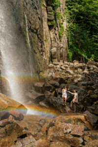 A couple stands on rocks in front of a double rainbow at a waterfall in the adirondack mountains.