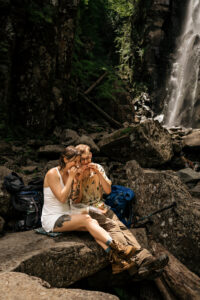 A couple eats sandwiches on a rock in front of a waterfall in the adirondack mountains.