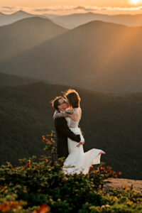 A couple who just eloped embrace on a high peak summit in the adirondack mountains at sunset and they are kissing while the bride is in the air.