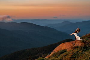 A couple who just eloped embrace on a high peak summit in the adirondack mountains at sunset and they are kissing while the bride is in the air.