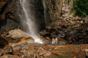 A couple stands on rocks in front of a double rainbow at a waterfall in the adirondack mountains.