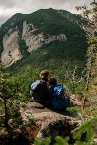 a couple leans on each other as they look at the view of a mountain with pine trees and backpacks on their backs.