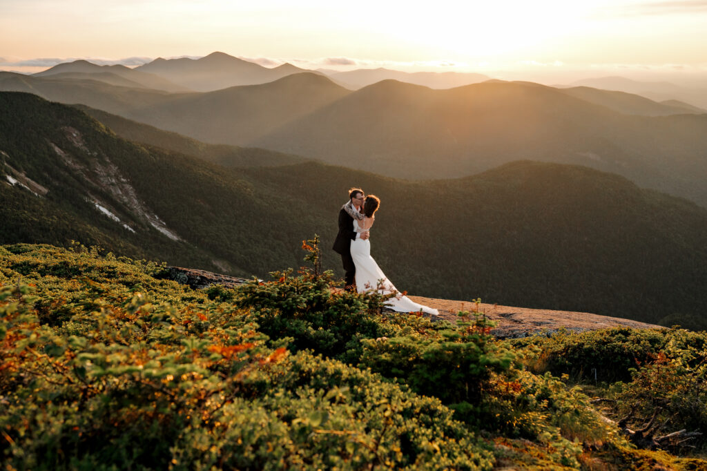 Couple in wedding attire kisses on top of a high peak in the Adirondack Mountains at sunset.