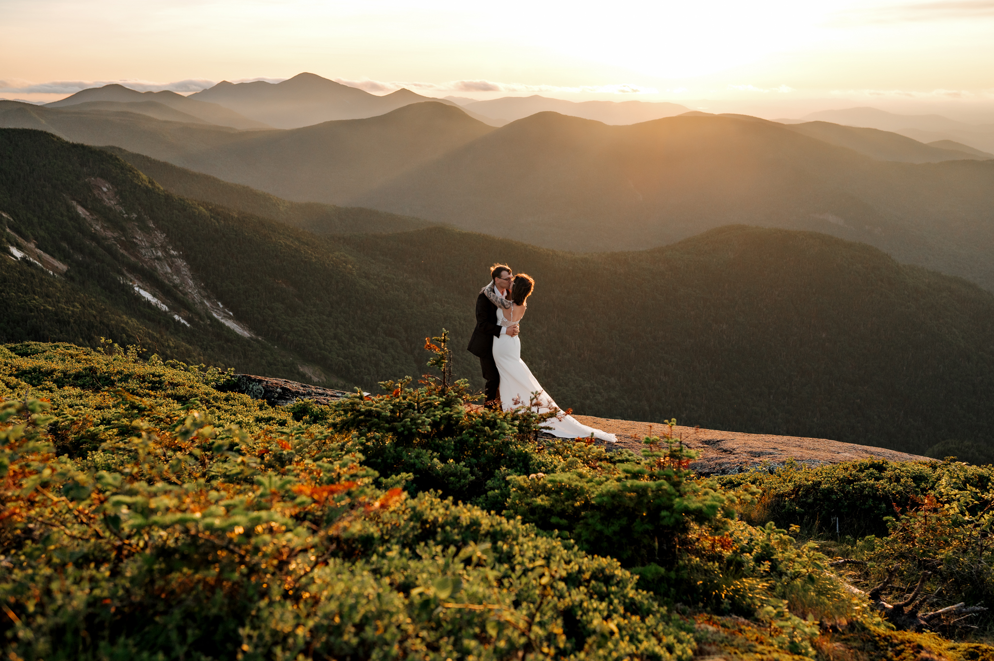 Couple in wedding attire kisses on top of a high peak in the Adirondack Mountains at sunset.