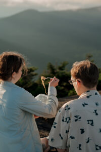 A couple eats ramen noodles at the top of a mountain in the Adirondacks with a mountain backdrop.