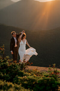 A couple dances on a high peak summit in the Adirondack Mountains with a mountain backdrop and golden sun rays shine on them as the brides dress flows in the wind.