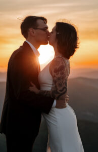 A couple kisses as the sun peaks through between them on top of a mountain in the adirondacks.