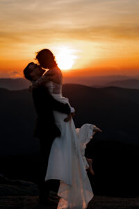 A groom lifts bride in the air on top of a high peak in the adirondack mountains at sunset and the picture shows the bride and groom silhouette and the sun setting behind them on the mountains.