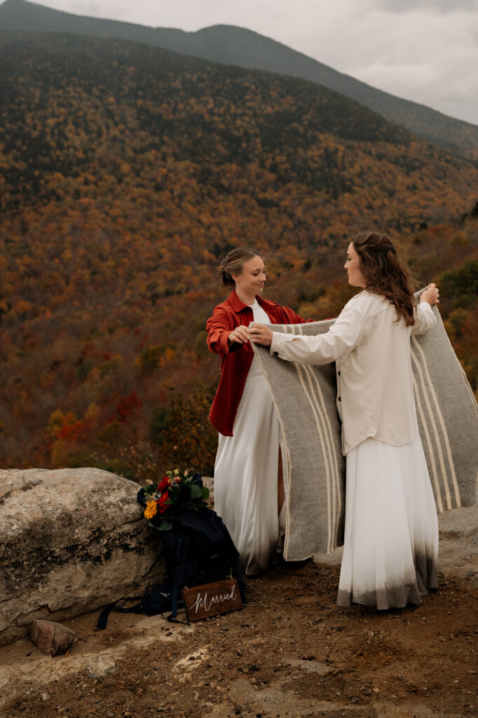 Two brides fold a picnic blanket and prepare to sit down and have hot cocoa.