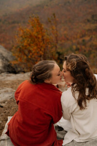 Two brides kissing with hot chocolate in their mugs and fall foliage behind them.
