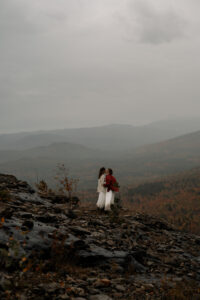 Two brides embrace at a cliff edge in a misty cloud overlooking mountains with fall foliage.