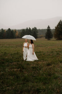 Two brides walk together in the rain with a clear umbrella in two white wedding dresses with a mountain backdrop in the Adirondacks.