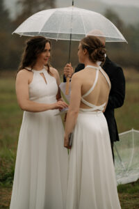 Two brides reading their vows under an umbrella in the rain.