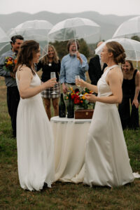 Two brides toast with eachother in their wedding dresses with their family behind them with umbrellas laughing.