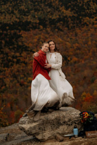 Two brides embrace each other on top of a rock with mountain foliage behind them as the wind blows their dresses.