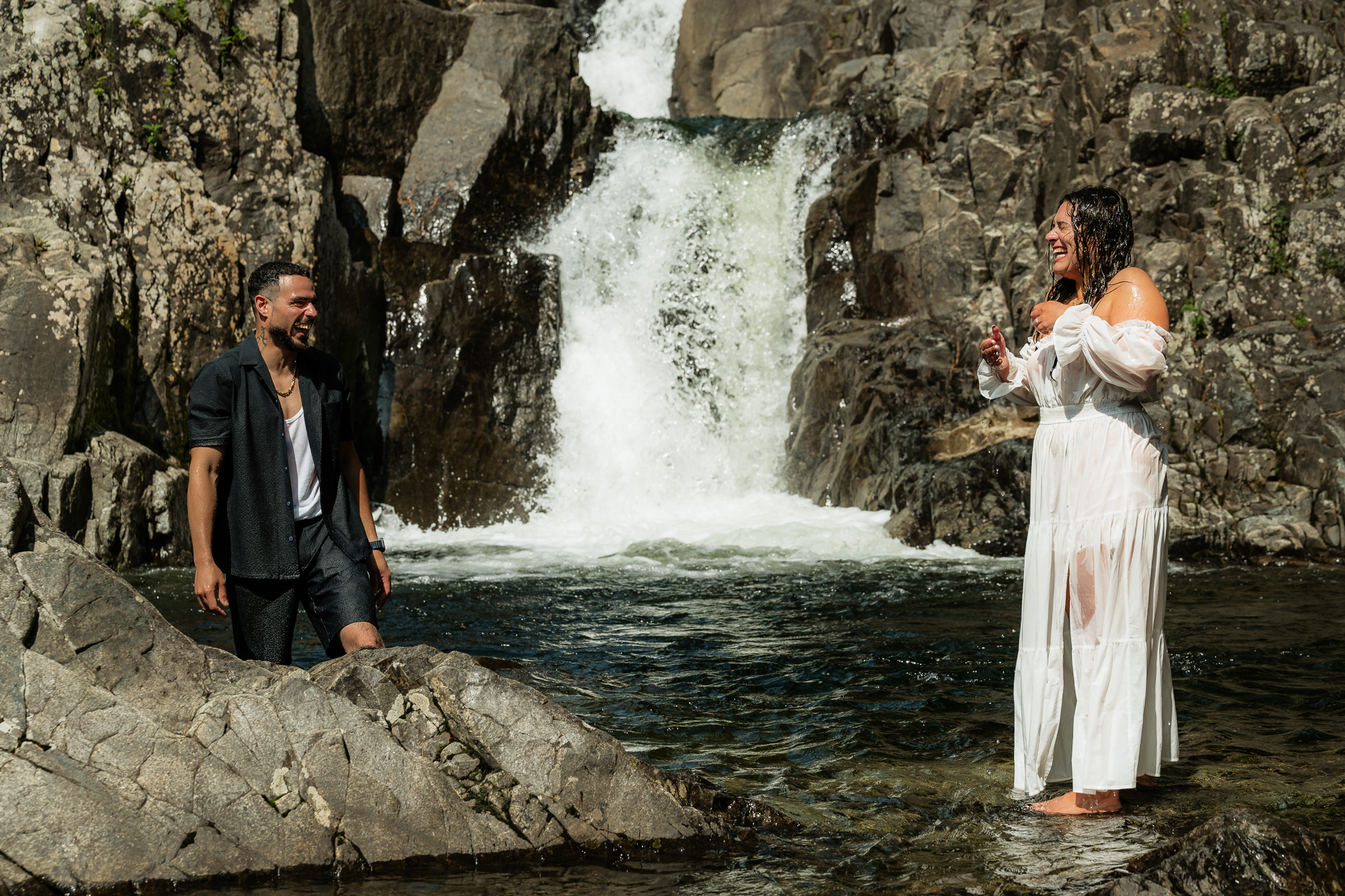 A couple who just eloped at a waterfall and jumped into the water in their wedding attire, splashing each other and laughing.