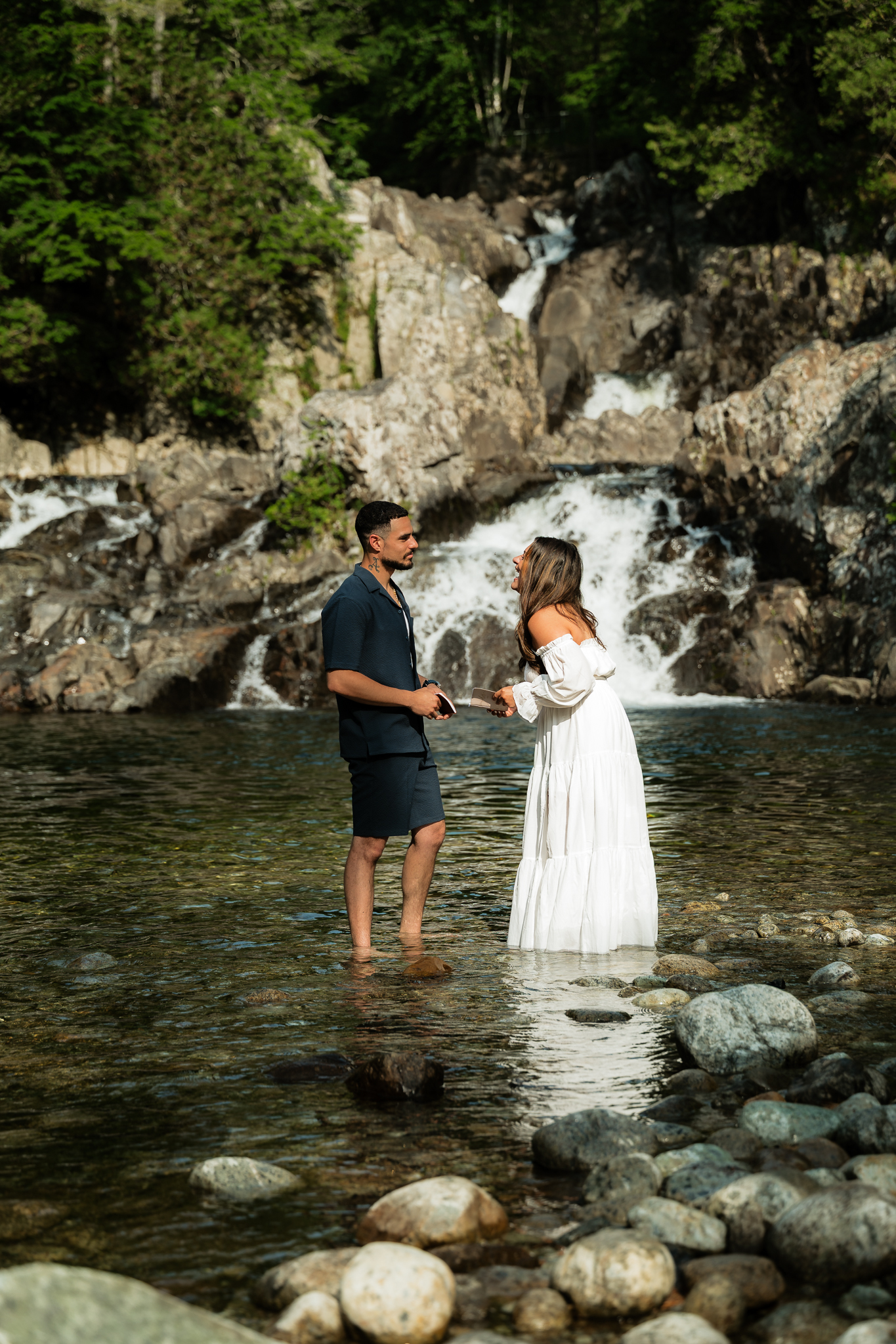 Couple stands at the base of a waterfall in the Adirondack Mountains reading their vows to each other laughing.