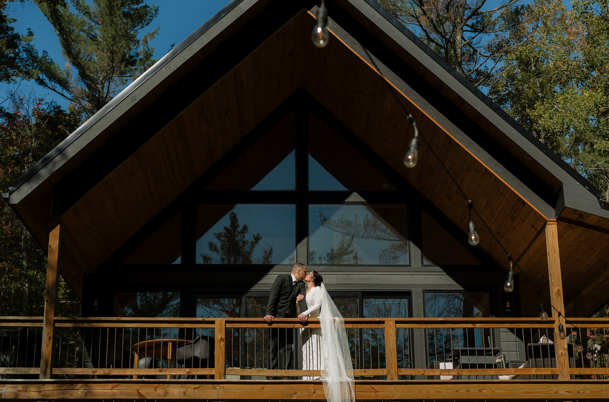 Couple on balcony of a frame cabin on sunny day kissing in wedding attire.