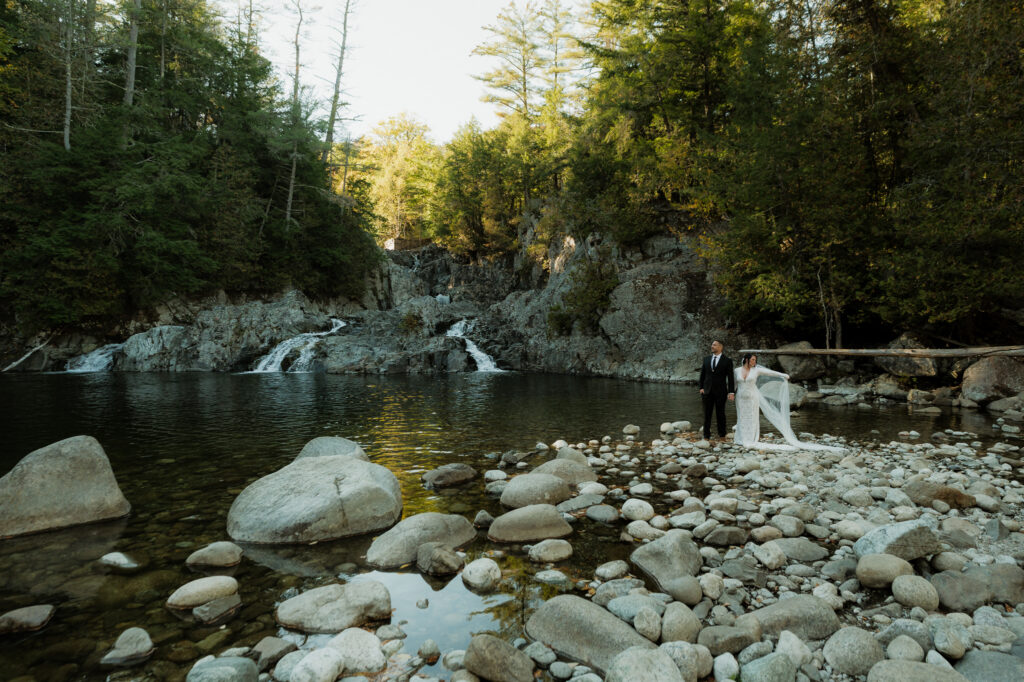 Couple stands at the edge of a waterfall on the rocks in wedding attire.