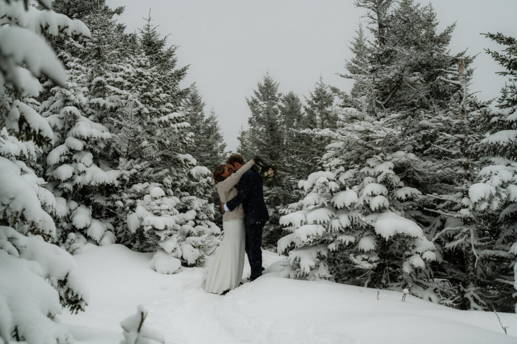 Couple hugs in a snowy forest clearing surrounded by pine in their wedding attire. 