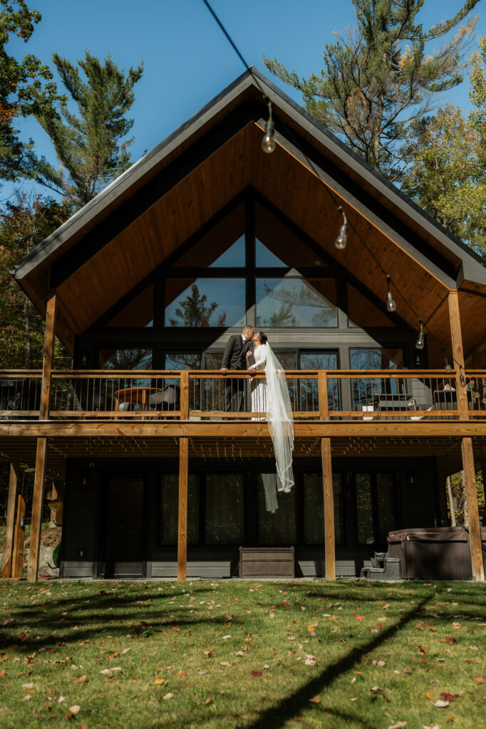 Bride and groom kiss on porch with brides veil draped over the ledge and framed by the a frame air bnb.
